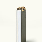 The Discbound Analog + 3 Refills is a vertically standing notebook with a dark cover and thick white pages, bound at the top with three brass disc binding rings, shown on a plain light background.