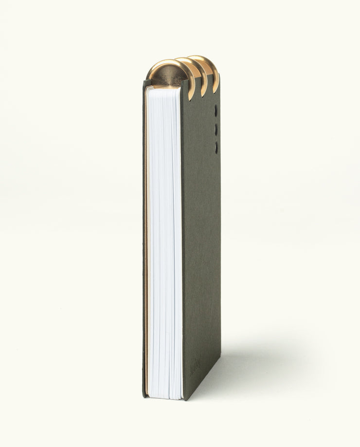 The Discbound Analog + 3 Refills is a vertically standing notebook with a dark cover and thick white pages, bound at the top with three brass disc binding rings, shown on a plain light background.