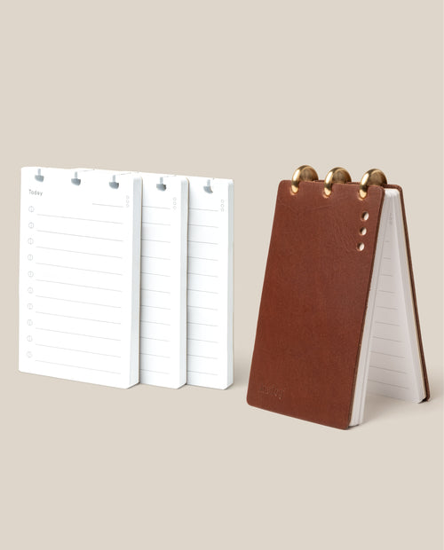 The Leather Discbound Analog + 3 Refills (Brown), a brown leather ring-bound notepad, stands upright beside three stacks of daily planner page refills on a light beige background.