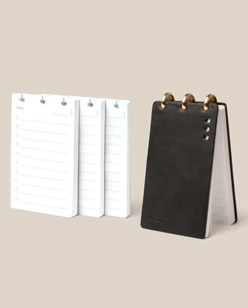 The Leather Discbound Analog + 3 Refills (Black) stands upright beside three stacks of “Today” daily planner pages with lines for notes and times, all displayed on a beige background—ideal for organization.