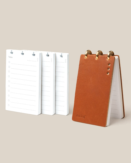 The Leather Discbound Analog + 3 Refills (Tan) features a tan full-grain leather notepad with gold disc rings, shown standing upright beside three stacks of loose refills on a beige background.