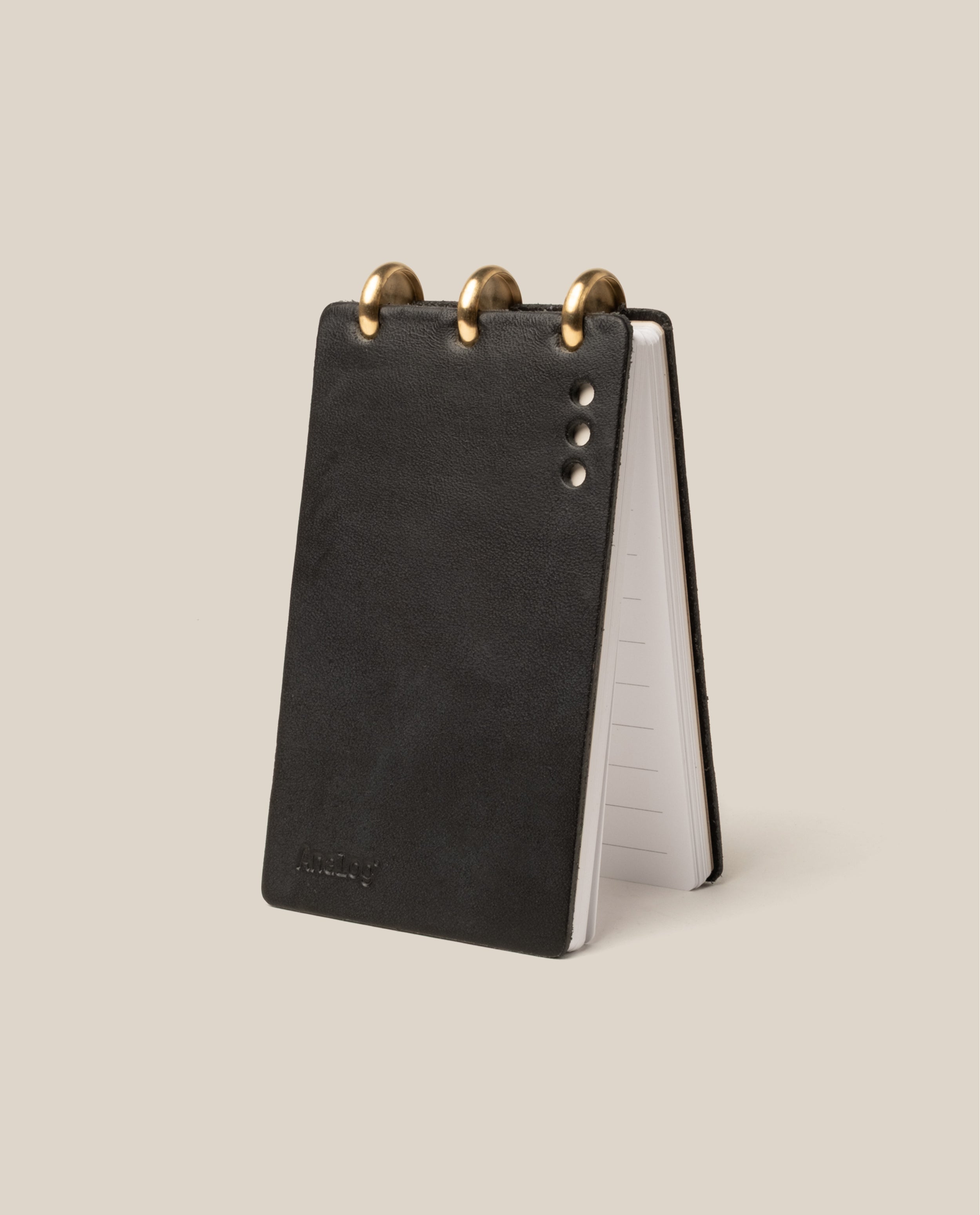 A compact Leather Discbound Analog (Black) notebook with three brass rings on top stands upright on a light beige background. Visible metal rivets and lined pages inside make it ideal for organizing Analog Task Cards.