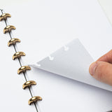 A hand lifts a dotted page in the Discbound Journal (Standard 6"x 8.5") with solid brass discs. The refillable journal rests open on a white surface, showing blank pages with light gray dots.