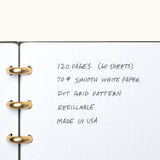 A close-up of the Discbound Journal (Standard 6"x 8.5") with solid brass discs, showing dotted grid pages and handwritten feature notes: 120 pages of 70# smooth white paper, refillable, dot grid pattern, made in USA.