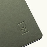 A close-up of the corner of the Discbound Journal (Standard 6"x 8.5") in green, featuring an embossed geometric logo on the cover and solid brass discs binding partially open white pages inside.