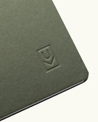 A close-up of the corner of an olive green Discbound Journal Bundle (8.5" x 11" Journal + 3 Refills) with a debossed logo featuring a U and a check mark inside a rectangle.