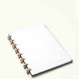 A Discbound Journal (Standard 6"x 8.5"), featuring a white dotted interior, black cover, and solid brass discs, rests on a light plain surface.