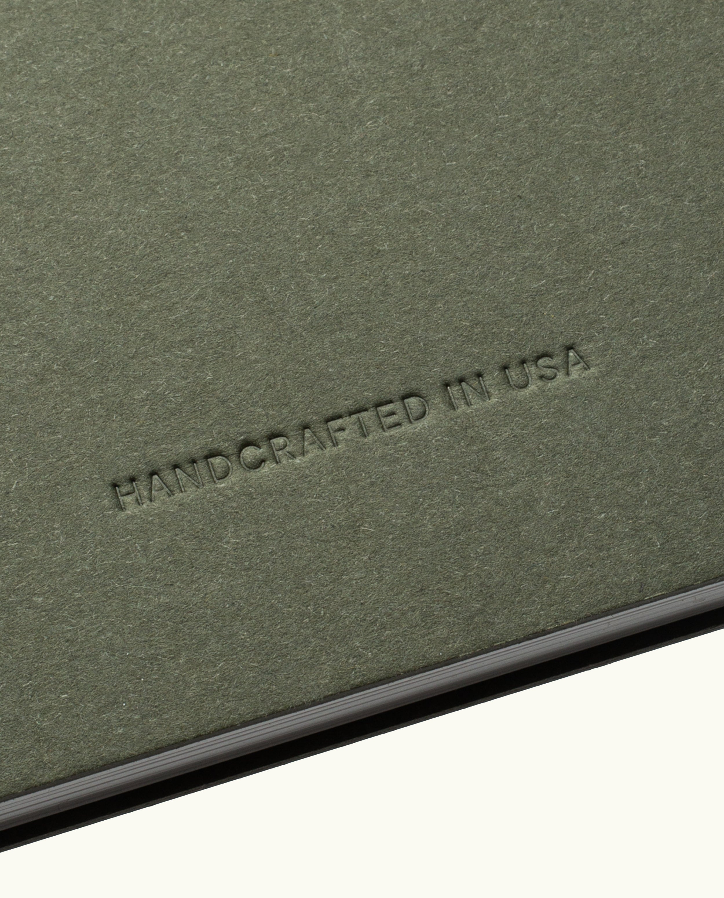 A close-up of a textured, olive-green surface with the words HANDCRAFTED IN USA embossed in uppercase letters. The lighting accentuates the rich texture and indentation of the text, reflecting the artisanal quality present in every Discbound Journal Bundle (8.5" x 11" Journal + 3 Refills)—including those featuring striking brass discs—all carefully crafted to be both refillable and distinctive.