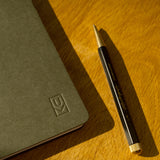 A black and gold pen rests on a wooden surface beside a closed, dark gray Discbound Journal (Standard 6"x 8.5") with an embossed logo in the corner, bathed in warm sunlight.