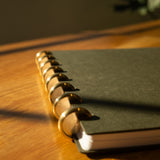 A close-up of the Discbound Journal (Standard 6"x 8.5") with solid brass discs, shown in dark colors on a wooden surface under warm lighting and soft shadows.