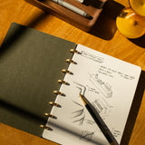 A Discbound Journal (Standard 6"x 8.5") with sketches and notes on architectural design sits open on a wooden desk, a black and gold pen resting on its refillable pages as sunlight casts shifting shadows across the scene.