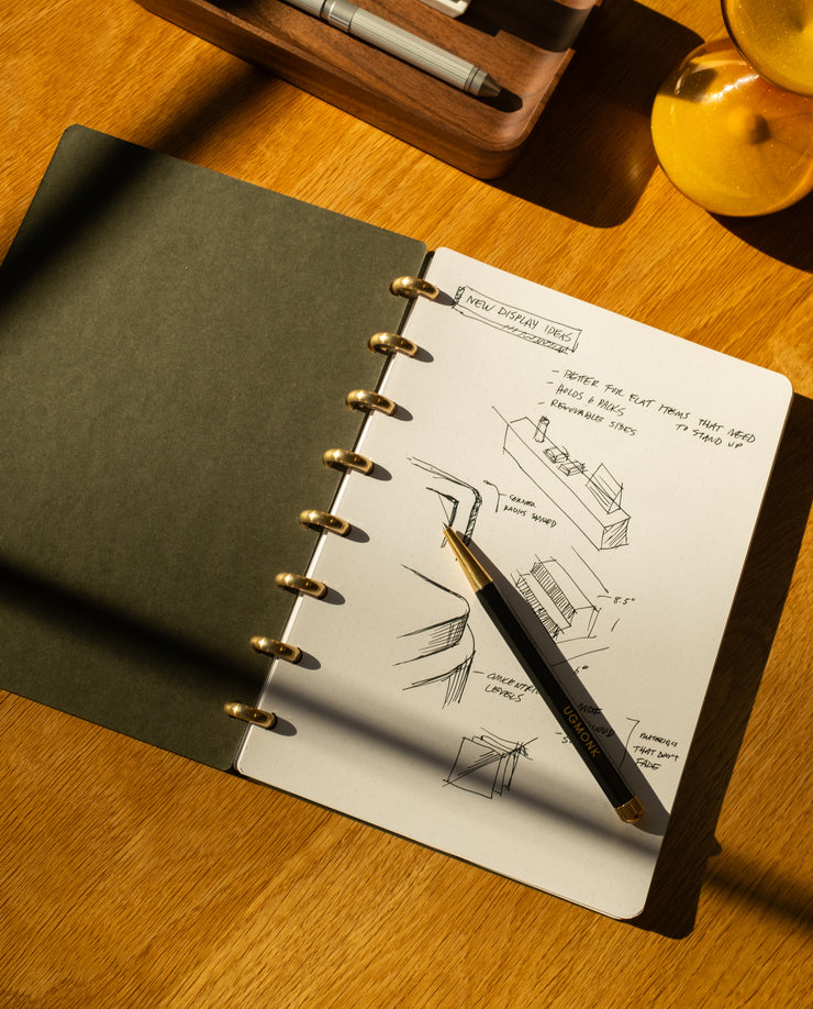 A Discbound Journal (Standard 6"x 8.5") with sketches and notes on architectural design sits open on a wooden desk, a black and gold pen resting on its refillable pages as sunlight casts shifting shadows across the scene.