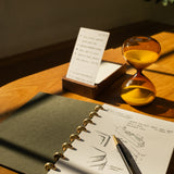 A Discbound Journal (Standard 6"x 8.5") with sketches and a pen rests on a wooden desk, next to an hourglass of yellow sand and an upright notepad with handwritten notes, all bathed in warm sunlight.