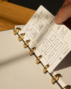A hand holds a small card with website wireframe sketches over an open Discbound Punch, its ring-bound dotted pages resting on a wooden desk, lit by sunlight.