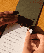 A person flips through a small discbound analog notebook with a to-do list, including tasks like reviewing a product, uploading products, and emailing copy. Sunlight casts shadows across the notepad and their hands as they prepare to add a Discbound Analog Refill (1-Pack) for their next tasks.