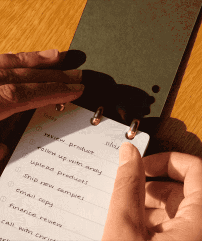 A person writes on the Discbound Analog + 3 Refills planner with brass disc binding, listing tasks such as reviewing and uploading products. Sunlight casts gentle shadows across the wooden surface beneath the planner.