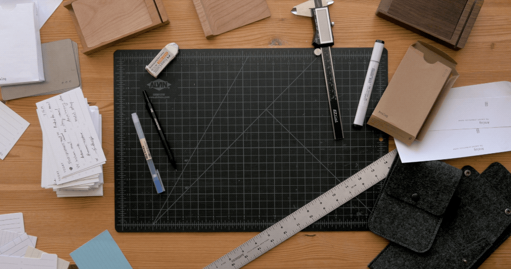 A black cutting mat sits on a wooden desk, surrounded by scattered index cards, notebooks, a ruler, pens, an eraser, a caliper, and a felt pouch, creating a workspace for writing or crafting.