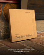 A brown cardboard box labeled UGMONK Analog Think Better on Paper leans against a brick wall, with the caption: The Analog Starter Kit (Black) includes everything you need to get started with the paper card system—task cards and an aluminum card stand—delivered right to your door.