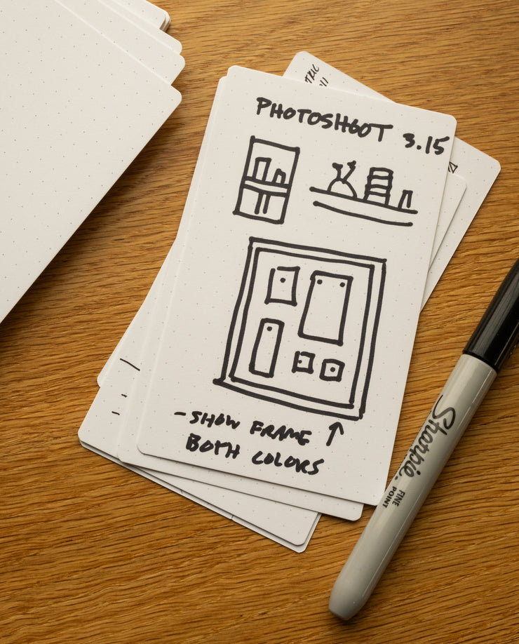 A stack of Analog Dot Grid Cards - Small (3-Pack) sits on a wooden surface, the top card filled with sketches and notes for Photoshoot 3.15: "show frame both colors." A black Sharpie rests nearby, ready for more ideas.