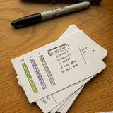 A pack of Analog Dot Grid Cards - Small (1-Pack) sits on a wooden table, the top card displaying a weekly workout plan and notes; ideal for analog planning. A black Sharpie marker rests nearby.