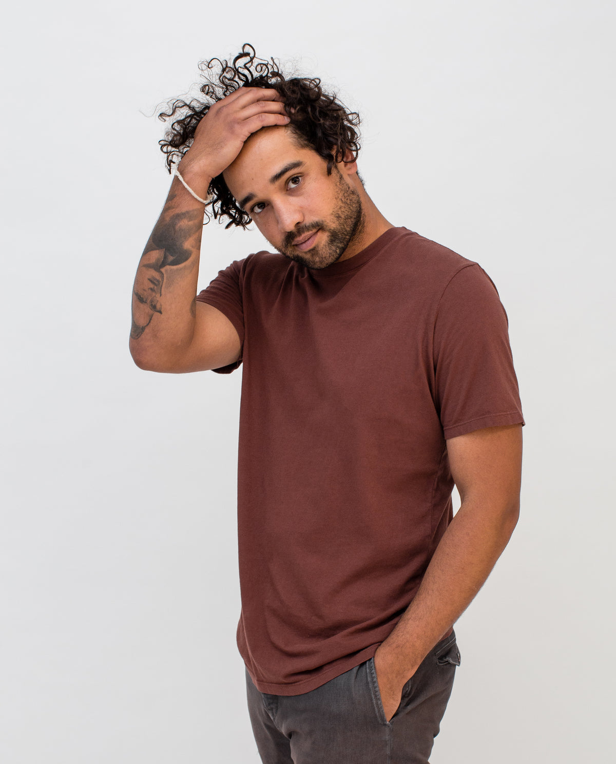 A man with curly hair and a beard, wearing a brown t-shirt and gray pants, poses against a plain white background with one hand in his pocket and the other touching his head.