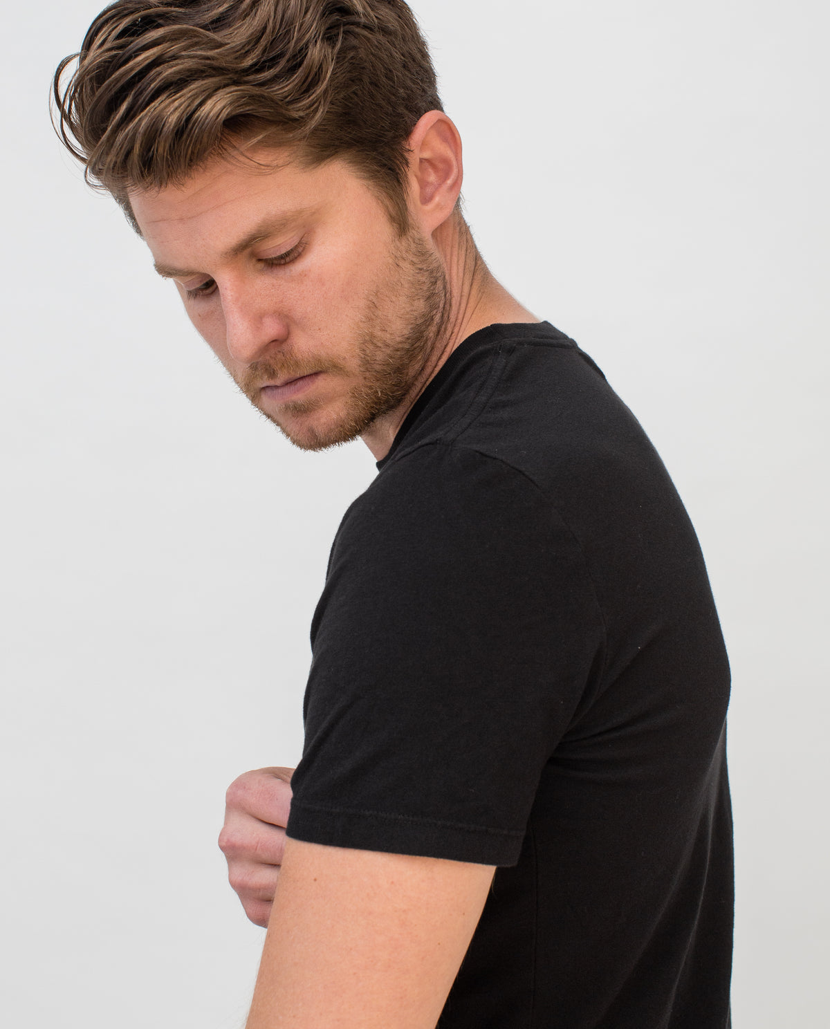 A man with light brown hair and a short beard, wearing a black t-shirt, looks down and slightly to his left against a plain white background.