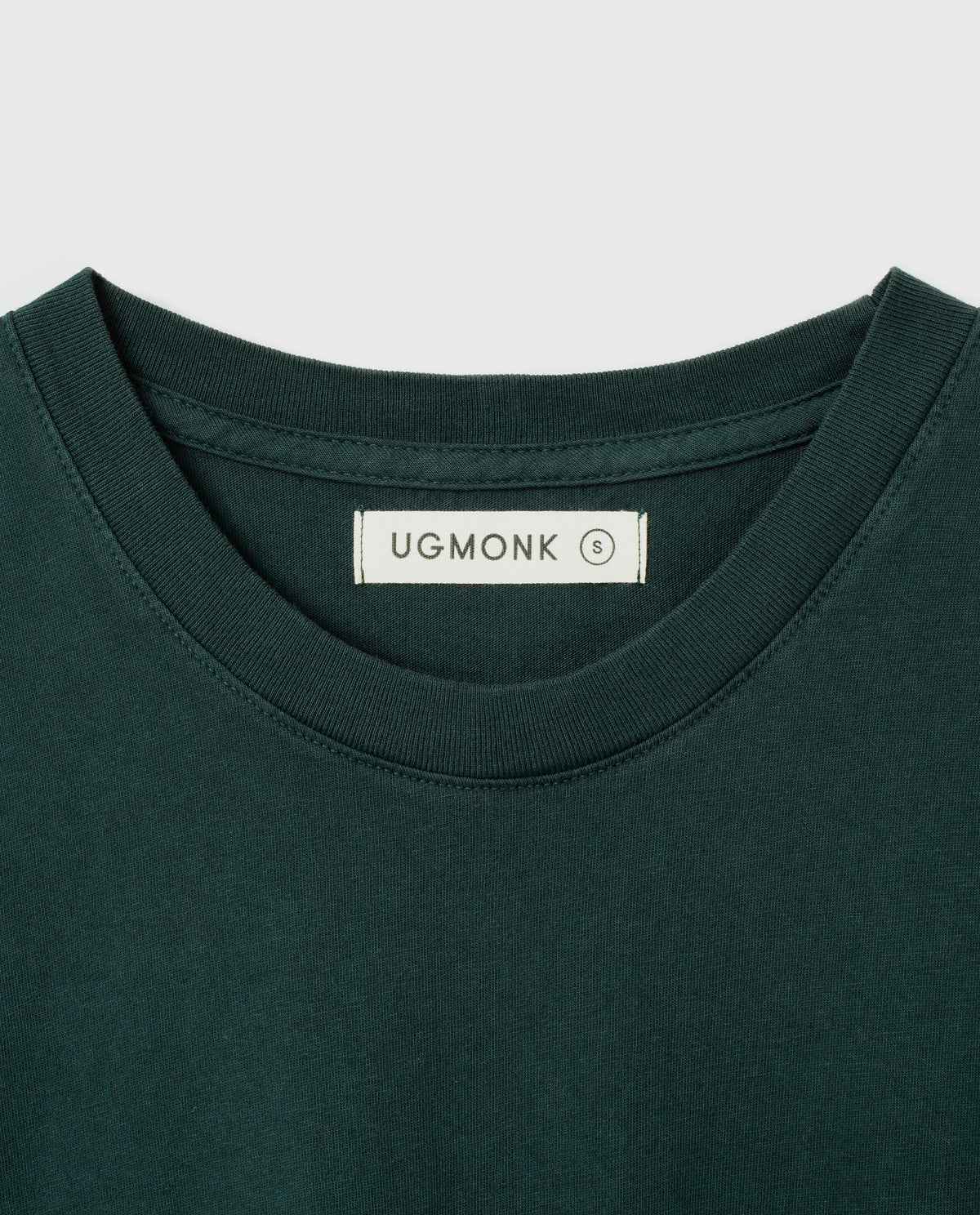 Dark green crewneck t-shirt with a white tag inside the collar displaying the brand name UGMONK and the size S. The shirt is neatly laid flat against a white background.