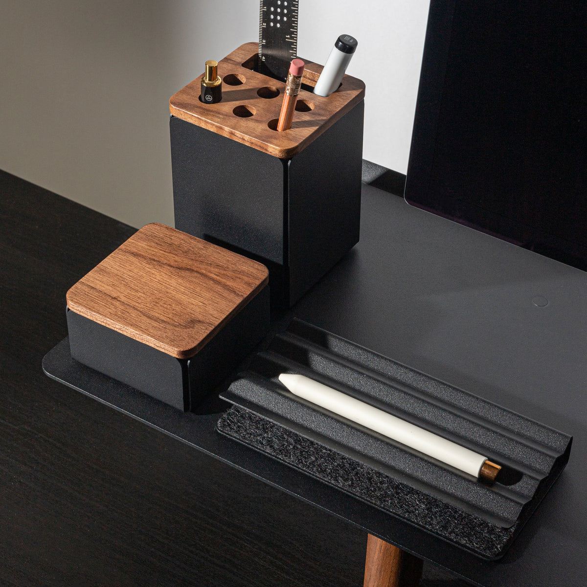 A modern desk organizer set with black and wood accents holds pens, a stylus, and small accessories next to a computer monitor on a dark desk. A closed box and an open tray with a white stylus are also visible.