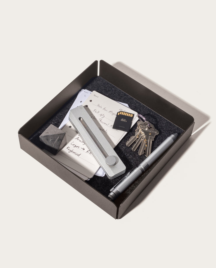 The Gather Large Tray (2x2 - Black) features a spacious, felt-like surface with a magnetic bottom, and holds items such as cards, a metal tool, a stone, an SD card, keyring keys, and two silver pens.
