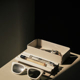 A pair of white sunglasses, a pen, and a small perfume bottle sit on the Gather Medium Tray (2x1 - White) next to an open cream-colored box, all highlighted by dramatic sunlight and shadows.