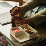 A person with painted nails opens a glass jar on a coffee table, revealing a box with a red playing card and leather strap atop the white Gather 2x2 Base Plate. A magazine and rug complete the cozy setting.