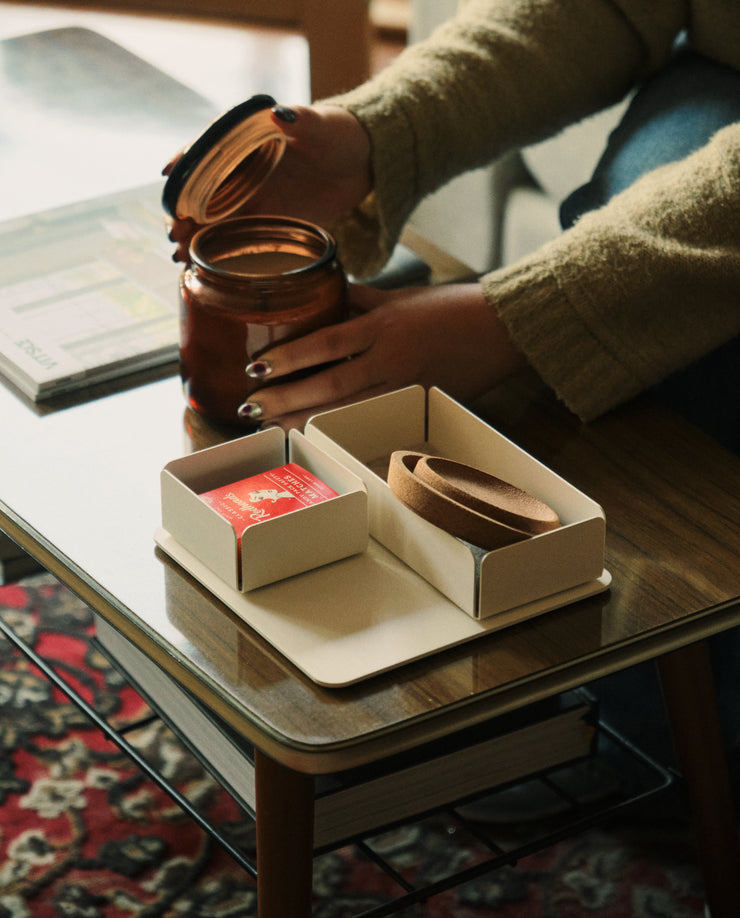 A person with painted nails opens a glass jar on a coffee table, revealing a box with a red playing card and leather strap atop the white Gather 2x2 Base Plate. A magazine and rug complete the cozy setting.