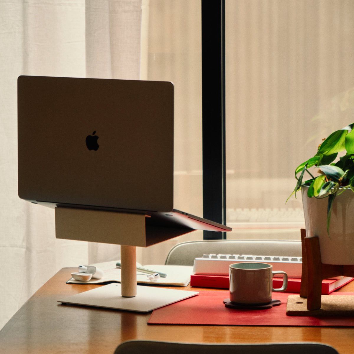 A laptop on a stand sits on a wooden desk with a keyboard, coffee cup, and potted plant near a window with natural light. The setup appears neat and organized, creating a cozy workspace atmosphere.