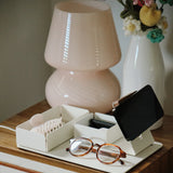 A pair of eyeglasses, a hair clip, and a small black wallet rest on the Gather Medium Tray (2x1 - White) atop a wooden nightstand with a striped drawer. A pink lamp and a white vase with crocheted flowers are in the background.