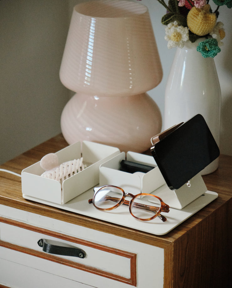 A pair of eyeglasses, a hair clip, and a small black wallet rest on the Gather Medium Tray (2x1 - White) atop a wooden nightstand with a striped drawer. A pink lamp and a white vase with crocheted flowers are in the background.