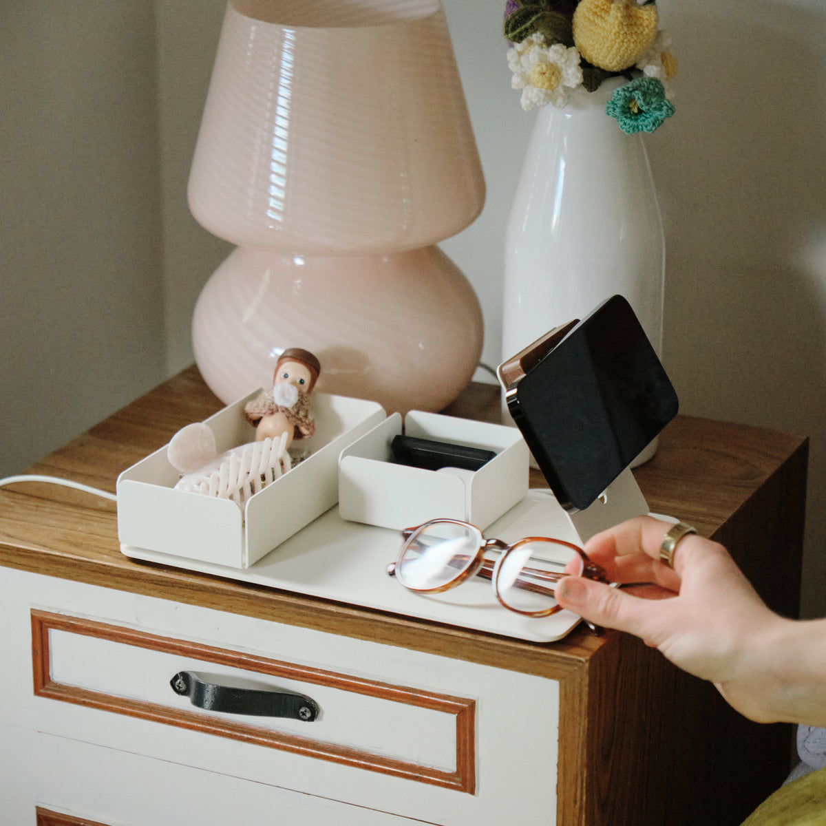 A hand picks up eyeglasses from a wooden nightstand with a pink lamp, a white vase of colorful flowers, a phone on a stand, and a small toy monkey in a tray.