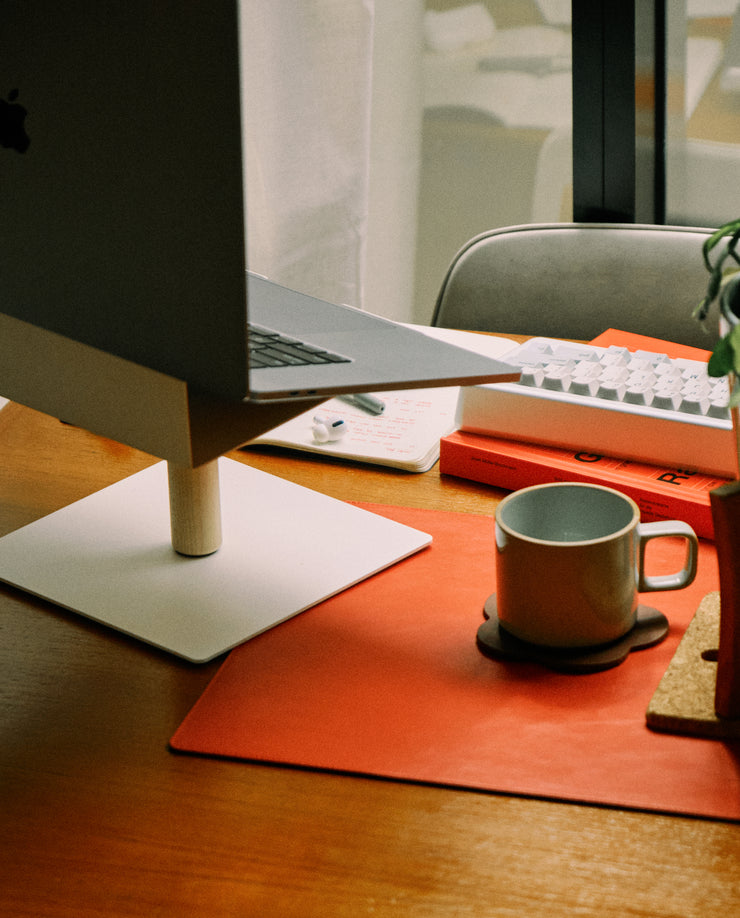 A workspace featuring a laptop on the Gather Laptop Stand (White/Maple), a notebook, pen, white keyboard, green mug on a coaster, and a potted plant on a desk with a red desk mat by the window.