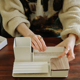 A person arranges the Gather Medium Tray (2x1 - White), a white steel desk organizer with wooden accents crafted in Pennsylvania, on a wooden table while wearing a beige fuzzy sweater and rings. The tray features compartments for organizing items.