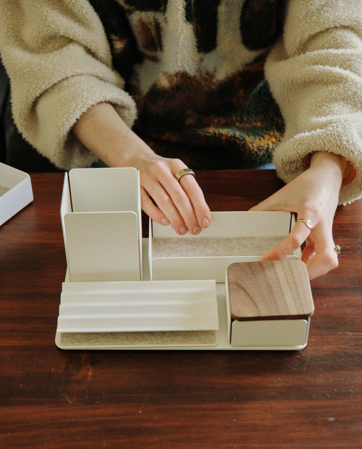 A person arranges the Gather Medium Tray (2x1 - White), a white steel desk organizer with wooden accents crafted in Pennsylvania, on a wooden table while wearing a beige fuzzy sweater and rings. The tray features compartments for organizing items.