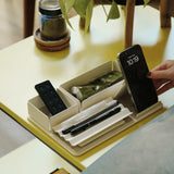 A hand picks up a smartphone from the Gather Medium Tray (2x1 - White), a powder coated steel desk organizer holding pens, a remote, and cream on a yellow table. In the background are a plant, cup, and books on a lower shelf.