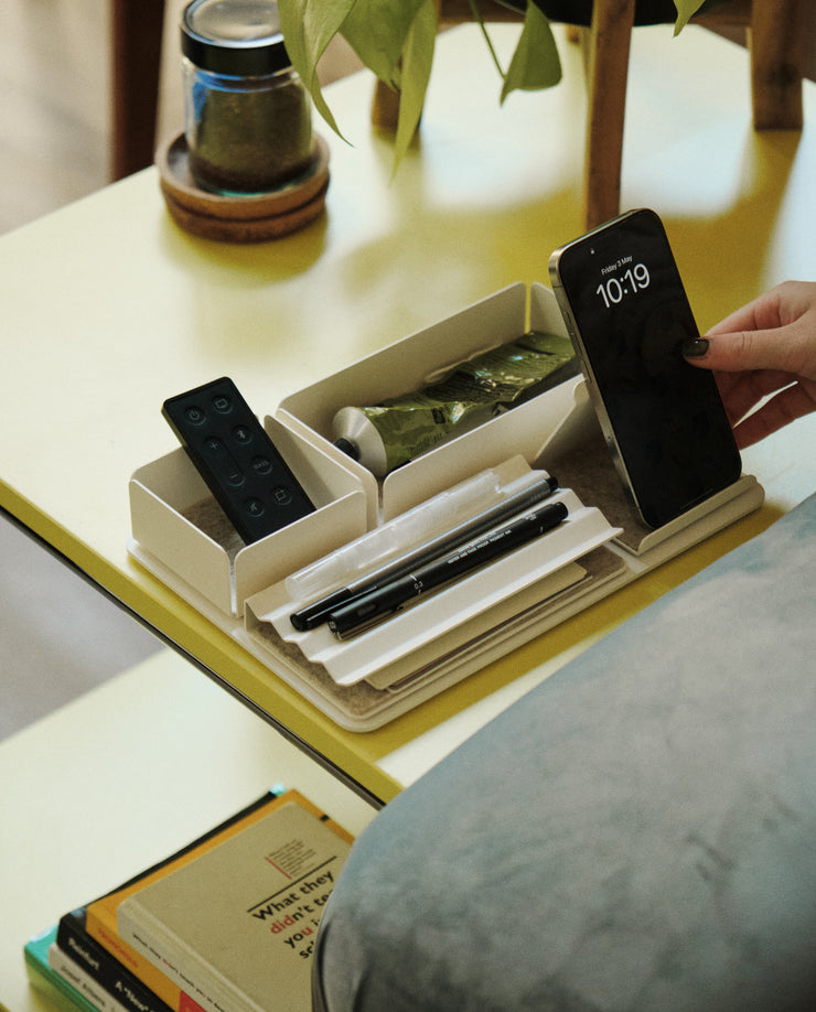 A person places a smartphone in the Gather Small Tray (1x1 - White), made in Pennsylvania, which also holds pens, a remote, and a green tube on a yellow table beside stacked books and a potted plant.
