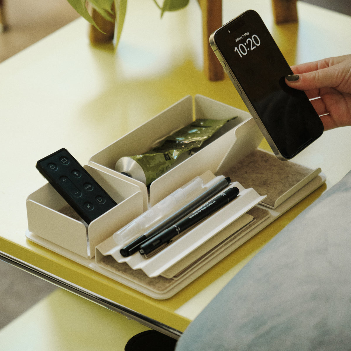 A hand holds a smartphone above an organized tray on a yellow table, containing pens, a remote control, and a small snack. A potted plant is in the background.