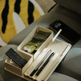 A Gather Medium Tray (2x1 - White), made of beige powder-coated steel, sits on a gray surface holding pens, a remote, a green tube, and other items. A smartphone shows 10:28 nearby, with part of a yellow, white, and black rug in the background.