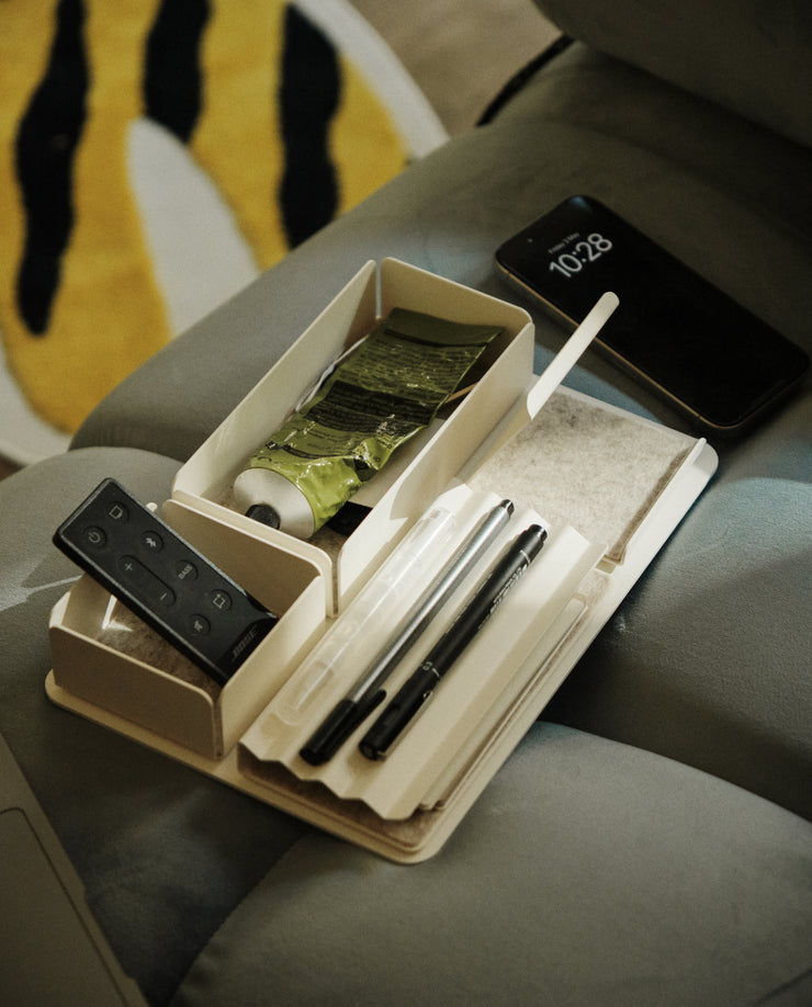 A Gather Medium Tray (2x1 - White), made of beige powder-coated steel, sits on a gray surface holding pens, a remote, a green tube, and other items. A smartphone shows 10:28 nearby, with part of a yellow, white, and black rug in the background.