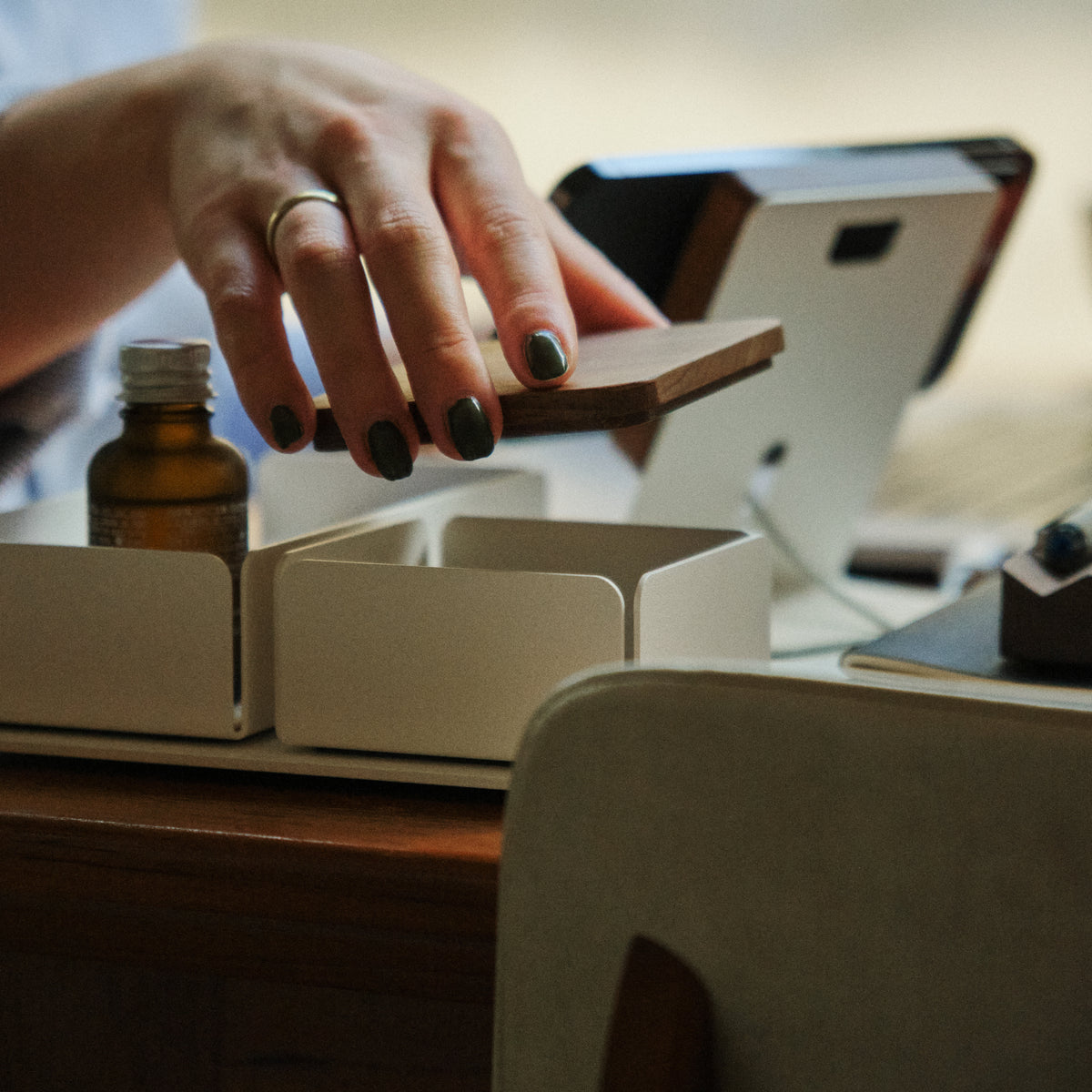 A hand with dark nail polish and a ring is reaching for a wooden lid on a white organizer, which also holds a small brown bottle, on a wooden table. A tablet device is visible in the background.