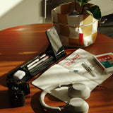 A wooden table featuring the Gather 1x4 Organizer Set (Black), a camera, headphones, smartphone, pens, an open notebook with notes, a plant in a fabric pot, and a box with Japanese writing—all bathed in warm sunlight.