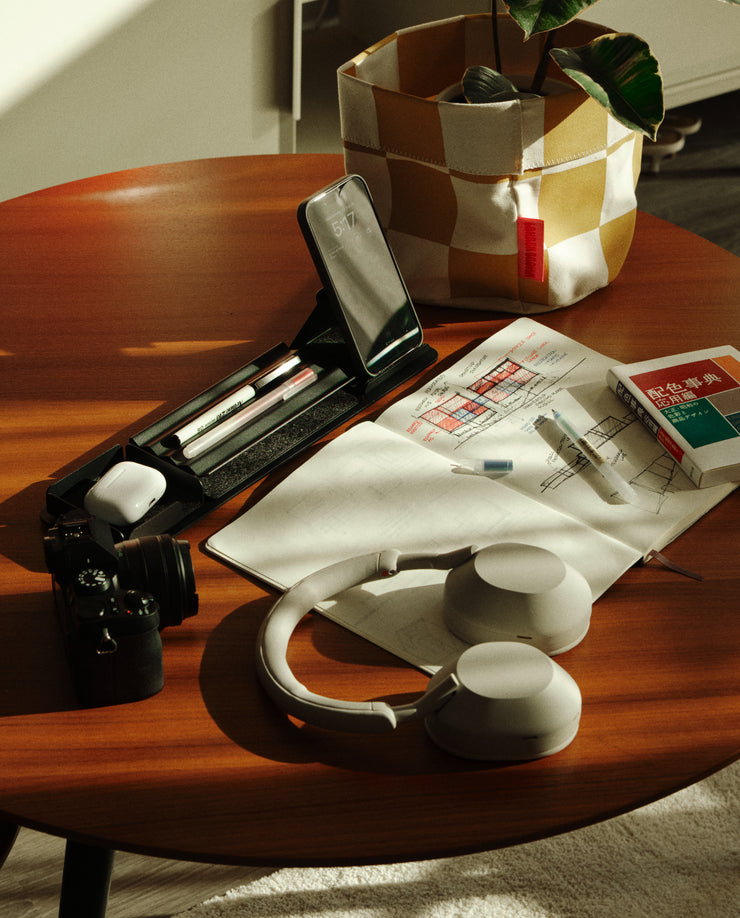 A wooden table featuring the Gather 1x4 Organizer Set (Black), a camera, headphones, smartphone, pens, an open notebook with notes, a plant in a fabric pot, and a box with Japanese writing—all bathed in warm sunlight.