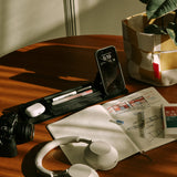 A wooden desk features headphones, a camera, pens, a smartphone on a stand, an open sketchbook, a small book, and magnetic Gather modules neatly arranged on the black Gather 1x4 Base Plate next to a checkered basket with a plant in sunlight.