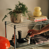 A metal wire shelf holds a potted plant, books, a yellow lamp, and skincare products. On the lower shelf, sunlight streams in as a hand reaches for a small jar near the Gather Large Tray (2x2 - Black) among folded clothes.