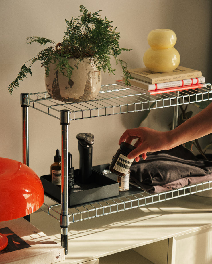 A metal wire shelf holds a potted plant, books, a yellow lamp, and skincare products. On the lower shelf, sunlight streams in as a hand reaches for a small jar near the Gather Large Tray (2x2 - Black) among folded clothes.